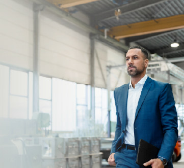 homem de barba vestindo um terno azul caminhando em um galpo de empresa carregando um tablet